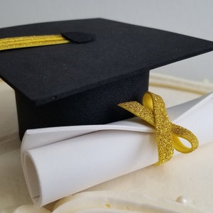May include: A black graduation cap with a gold tassel and a rolled-up white diploma tied with a gold ribbon. The items are placed on a white surface, likely a cake, with decorative frosting and small pearl-like accents.