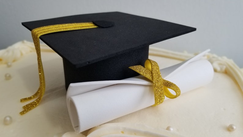 May include: A black graduation cap with a gold tassel and a rolled-up white diploma tied with a gold ribbon. The items are placed on a white surface, likely a cake, with decorative frosting and small pearl-like accents.