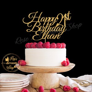 Pode incluir: Topper de bolo de glitter dourado com o texto "Happy 1st Birthday Ethan". O topper est&aacute; em um bolo branco com framboesas no topo. O bolo est&aacute; em um suporte de bolo de madeira.