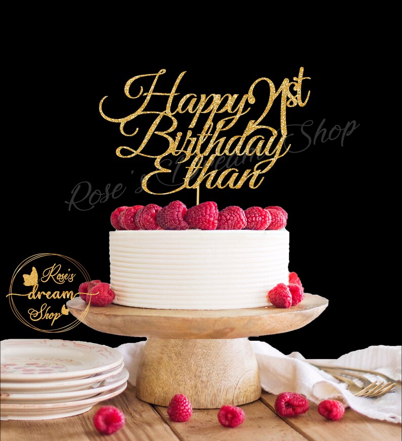 Pode incluir: Topper de bolo de glitter dourado com o texto "Happy 1st Birthday Ethan". O topper est&aacute; em um bolo branco com framboesas no topo. O bolo est&aacute; em um suporte de bolo de madeira.