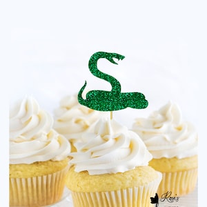 以下が含まれることがあります： 緑色のグリッターのスネークのカップケーキトッパー。ヘビは頭を上げて丸まっています。トッパーはつまようじに付いており、カップケーキに置く準備ができています。