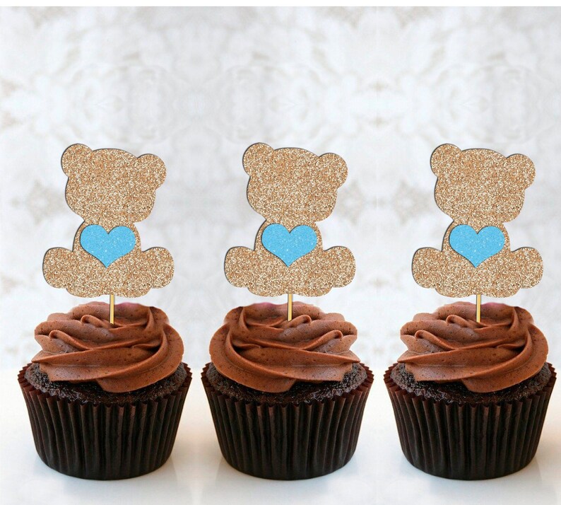 May include: Three chocolate cupcakes with gold glitter teddy bear toppers holding blue hearts. The cupcakes are frosted with chocolate frosting.