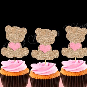 May include: Three cupcake toppers featuring a gold glitter teddy bear holding a pink heart. The toppers are on top of cupcakes with pink frosting.