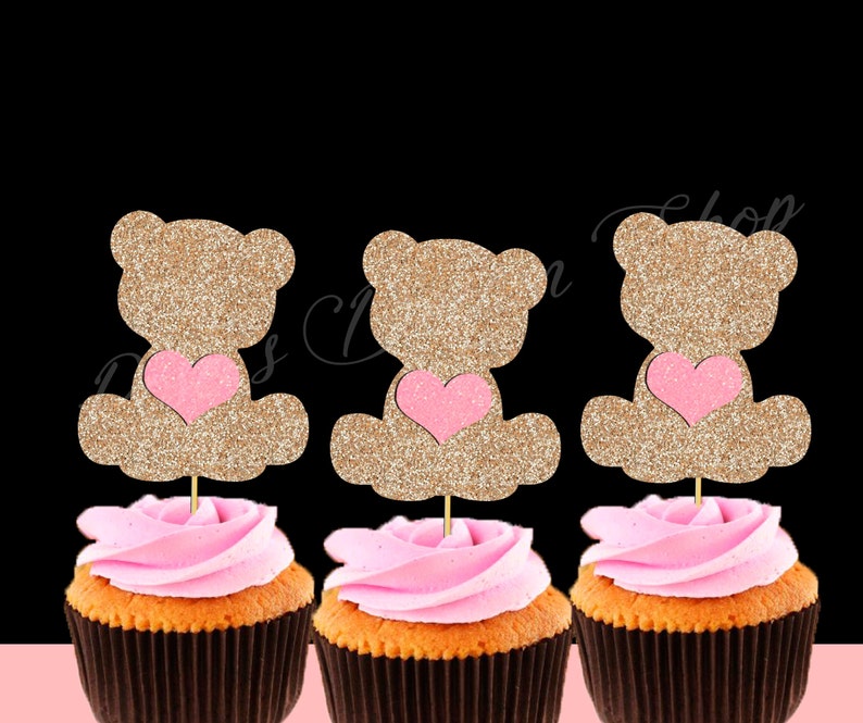 May include: Three cupcake toppers featuring a gold glitter teddy bear holding a pink heart. The toppers are on top of cupcakes with pink frosting.