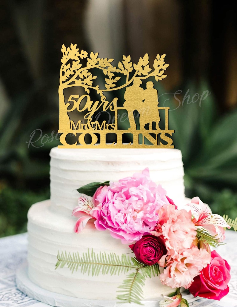 May include: A two-tiered white wedding cake decorated with pink and white flowers. A gold cake topper features a couple sitting on a bench under a tree with the text "50 yrs Mr & Mrs COLLINS"