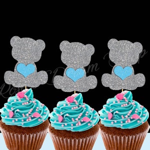 May include: Three cupcakes decorated with blue frosting and pink sprinkles, each topped with a silver glitter bear cupcake topper holding a blue heart.