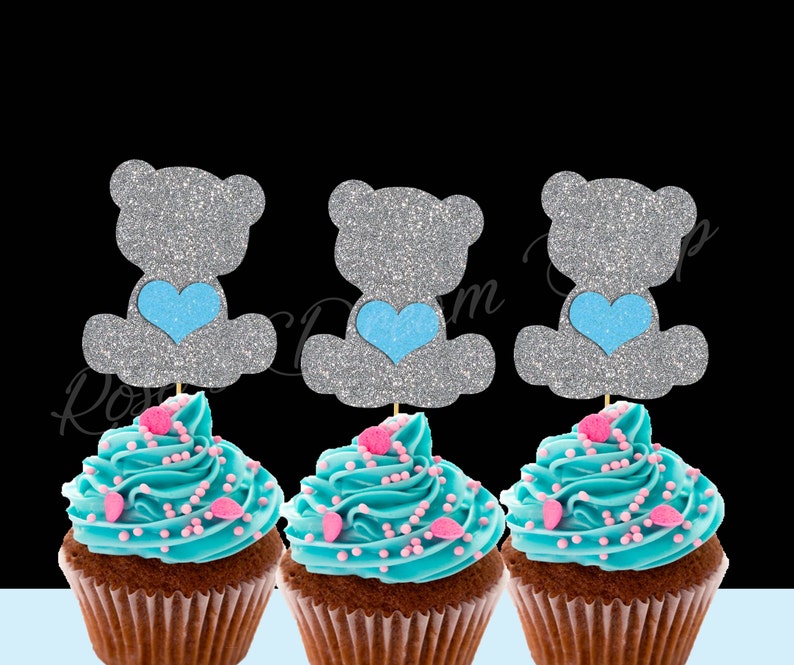 May include: Three cupcakes decorated with blue frosting and pink sprinkles, each topped with a silver glitter bear cupcake topper holding a blue heart.