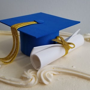 May include: A decorated cake with a blue graduation cap and a rolled diploma. The cap has a gold tassel, and the diploma is tied with a gold ribbon. The cake is white with decorative frosting and small pearl-like sprinkles.
