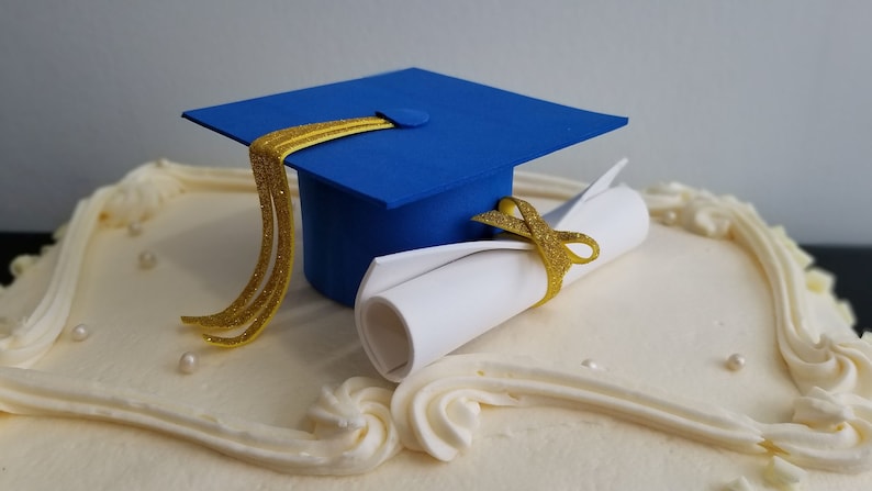 May include: A decorated cake with a blue graduation cap and a rolled diploma. The cap has a gold tassel, and the diploma is tied with a gold ribbon. The cake is white with decorative frosting and small pearl-like sprinkles.