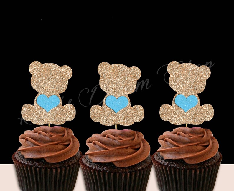 May include: Three chocolate cupcakes with brown frosting topped with glitter teddy bear cupcake toppers. Each teddy bear is holding a light blue heart.