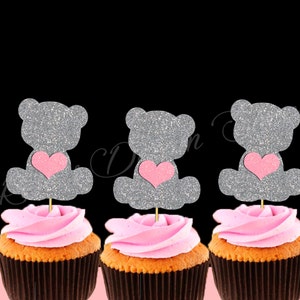 May include: Three cupcakes with pink frosting and silver glitter teddy bear toppers with pink hearts. The teddy bears are holding the hearts in their paws.