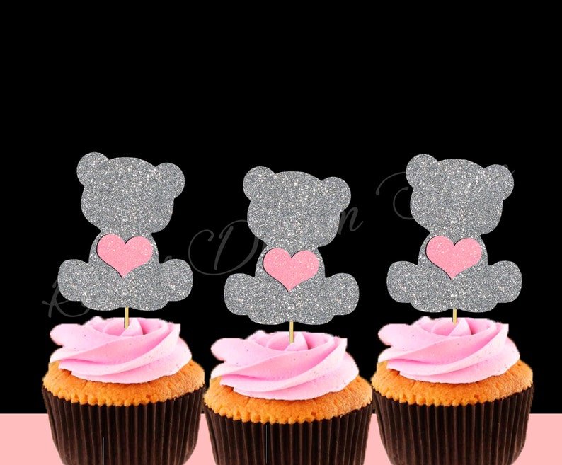 May include: Three cupcakes with pink frosting and silver glitter teddy bear toppers with pink hearts. The teddy bears are holding the hearts in their paws.