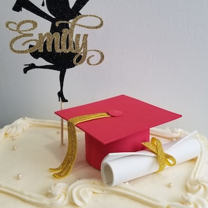 May include: A graduation cake with white frosting, decorated with a red mortarboard, a rolled diploma, and a black and gold glitter topper with the name "Emily". The cake is a celebratory dessert.