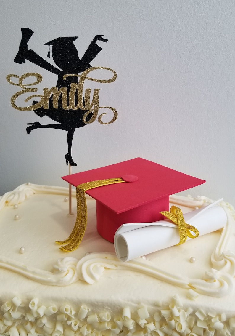 May include: A graduation cake with white frosting, decorated with a red mortarboard, a rolled diploma, and a black and gold glitter topper with the name "Emily". The cake is a celebratory dessert.