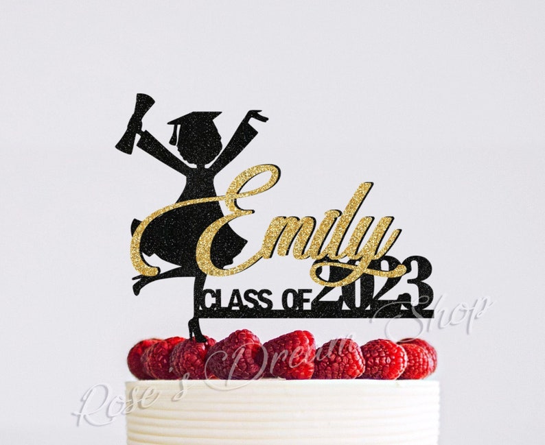 May include: Black silhouette of a graduate with a gold glitter name "Emily" and the text "Class of 2023" on a white cake with red raspberries.