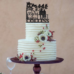 May include: A two-tiered white wedding cake with a black silhouette cake topper of a couple sitting on a bench under a tree. The topper says "50 yrs Mr & Mrs COLLINS". The cake is decorated with white anemones and red berries.