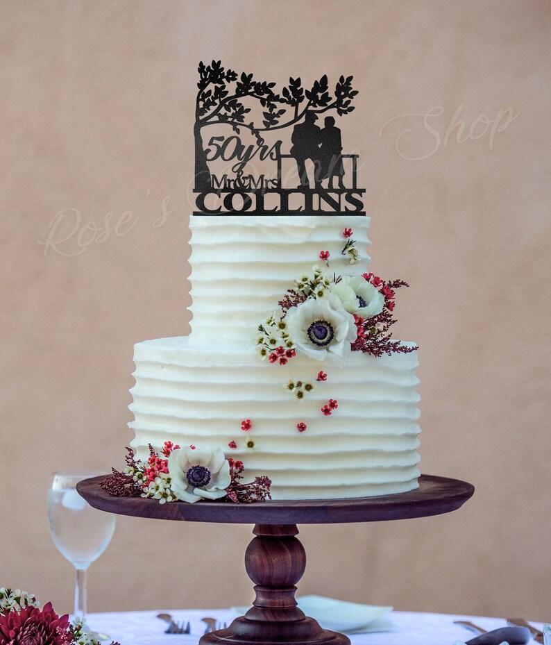 May include: A two-tiered white wedding cake with a black silhouette cake topper of a couple sitting on a bench under a tree. The topper says "50 yrs Mr & Mrs COLLINS". The cake is decorated with white anemones and red berries.