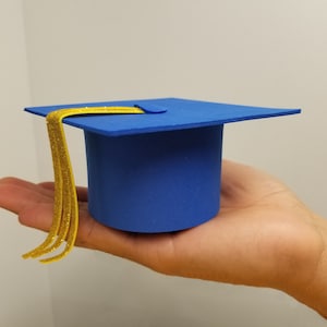 May include: A miniature blue graduation cap with a gold tassel. The cap is held in a hand, showcasing its small size and the details of the design. The cap's square top and cylindrical base are clearly visible.
