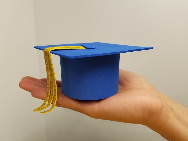 May include: A miniature blue graduation cap with a gold tassel. The cap is held in a hand, showcasing its small size and the details of the design. The cap's square top and cylindrical base are clearly visible.