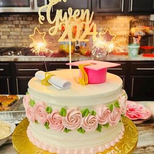 May include: A decorated graduation cake with pink roses and green leaves. The cake has a gold glitter topper that says "Gwen 2024". A pink graduation cap and a rolled diploma are on top. Sparklers are lit.