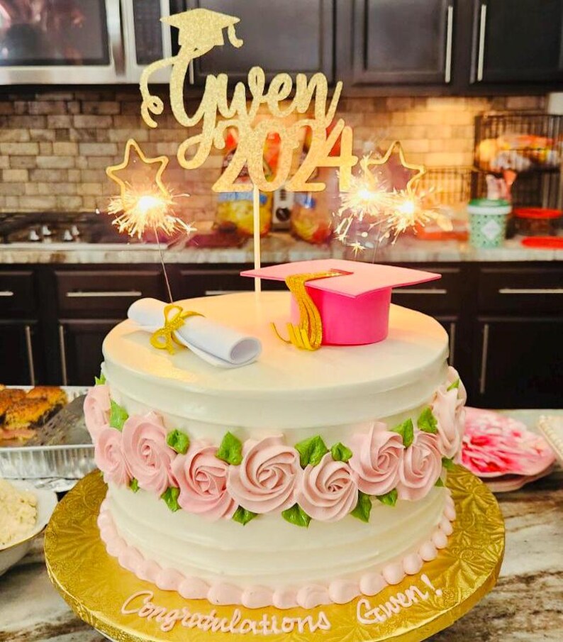 May include: A decorated graduation cake with pink roses and green leaves. The cake has a gold glitter topper that says "Gwen 2024". A pink graduation cap and a rolled diploma are on top. Sparklers are lit.