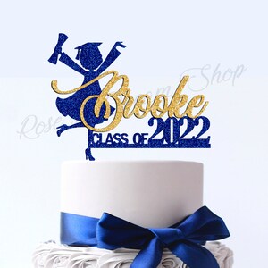 May include: Blue and gold glitter graduation cake topper with a silhouette of a female graduate, the name "Brooke" and the text "Class of 2022".