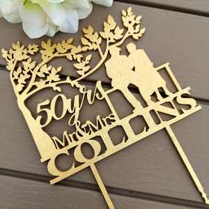 May include: Gold-colored wooden cake topper with a silhouette of a couple standing under a tree. The topper says "50 yrs Mr & Mrs Collins".