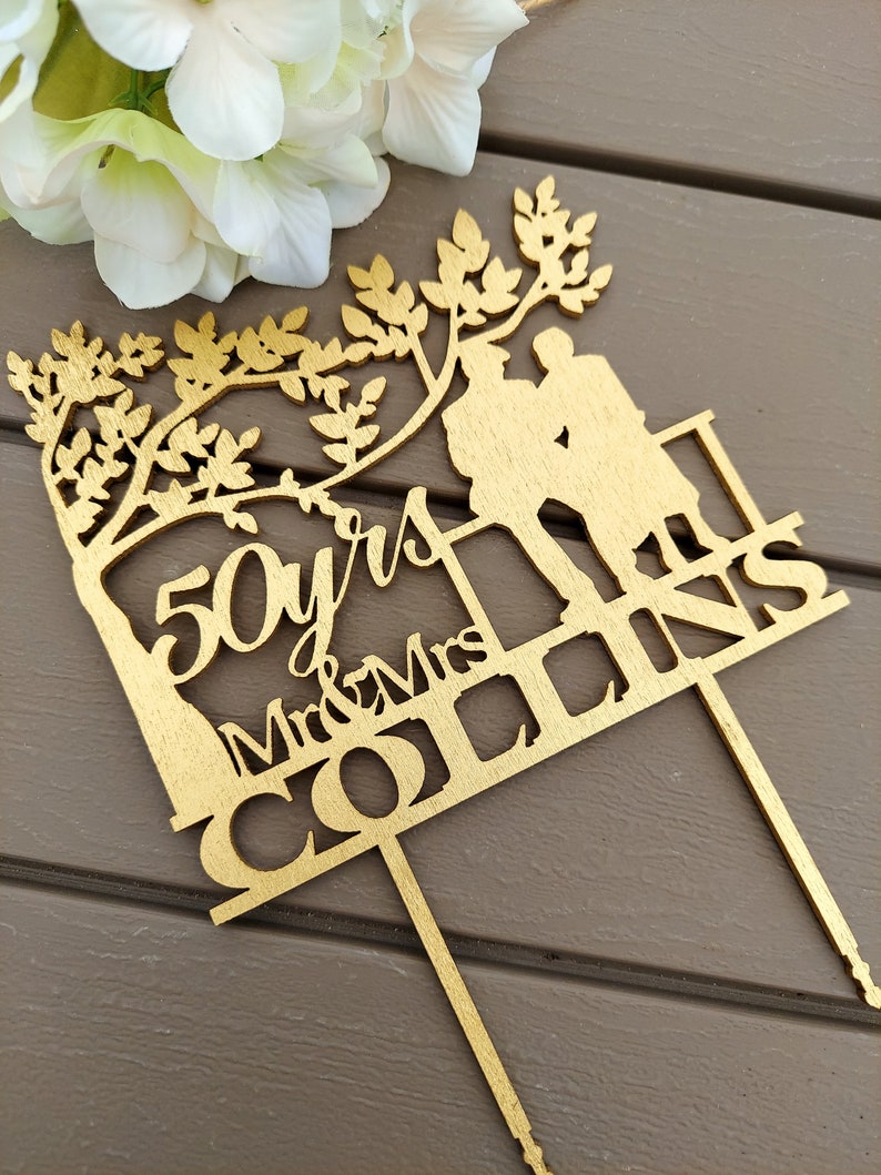 May include: Gold-colored wooden cake topper with a silhouette of a couple standing under a tree. The topper says "50 yrs Mr & Mrs Collins".
