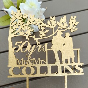 May include: A wooden cake topper with a silhouette of a couple standing under a tree. The topper says "50 yrs Mr & Mrs COLLINS".