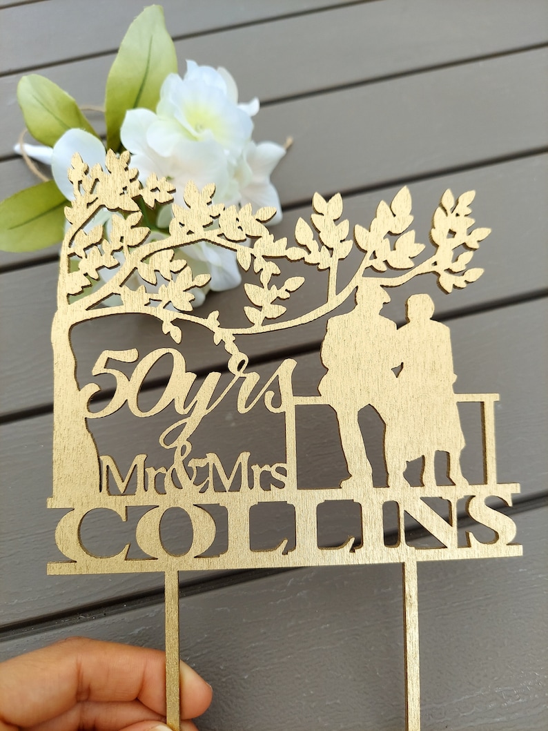 May include: A wooden cake topper with a silhouette of a couple standing under a tree. The topper says "50 yrs Mr & Mrs COLLINS".