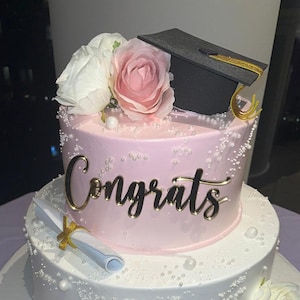 May include: A two-tiered graduation cake with pink frosting, adorned with white and pink roses, and a black graduation cap. The cake is decorated with gold script that says "Congrats" and a rolled diploma.
