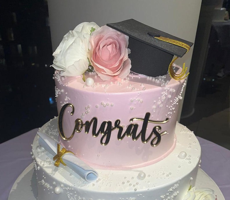 May include: A two-tiered graduation cake with pink frosting, adorned with white and pink roses, and a black graduation cap. The cake is decorated with gold script that says "Congrats" and a rolled diploma.