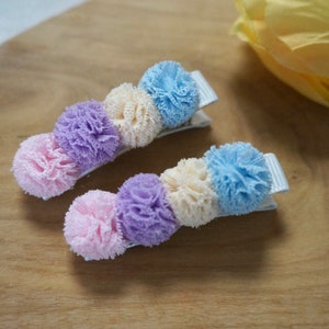 Puede incluir: Dos juegos de pinzas para el pelo con pompones de colores pastel. Los pompones son en tonos de rosa, morado, crema y azul.