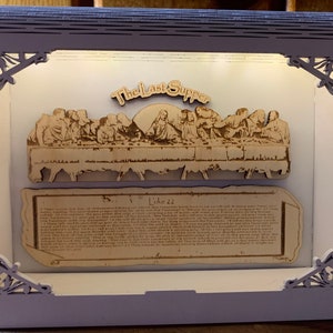 Op de afbeelding: Een houten ingelijst kunstwerk dat het Laatste Avondmaal afbeeldt met een passage uit Lukas 22. Het kunstwerk wordt van achteren verlicht, waardoor een warme gloed op de scène valt. De lijst is lichtpaars met ingewikkelde decoratieve hoeken.