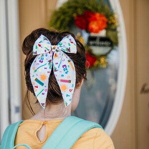 May include: A young person with brown hair wearing a yellow shirt and a green and white floral rucksack. The person has a white bow in their hair with a colourful pattern of letters, numbers, and school supplies.