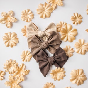May include: Three brown fabric hair bows with a satin finish. The bows are arranged in a stack, with the largest bow at the bottom and the smallest bow at the top. The bows are surrounded by small gold paper flowers.