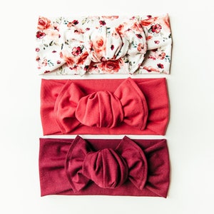 May include: Three headbands in different colours and patterns. The top headband is white with a floral print. The middle headband is a solid coral colour. The bottom headband is a solid burgundy colour. All three headbands are tied in a bow.