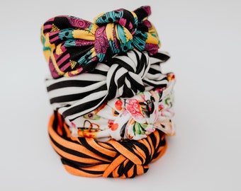 May include: A stack of four colorful fabric headbands with knotted bows. The headbands feature different patterns, including stripes, floral designs, and a Halloween-themed print.