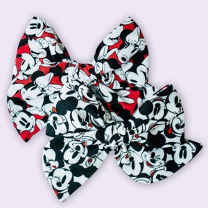 Peut inclure: Deux pinces à cheveux en forme de nœud rouge et blanc avec un imprimé Mickey Mouse noir et blanc.