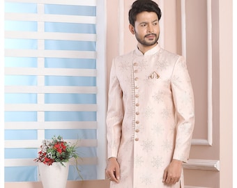 Puede incluir: Una kurta de hombre de color rosa claro con un estampado floral blanco y botones dorados. La kurta se lleva con pantalones blancos y zapatos blancos.