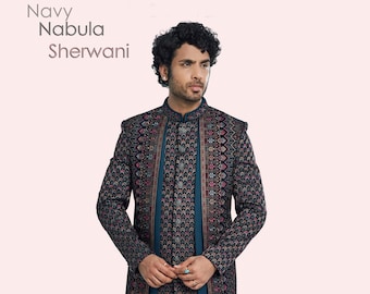 Puede incluir: Un sherwani azul marino estampado con un frente largo y abierto y una camiseta interior azul turquesa. El sherwani tiene un patrón geométrico con detalles dorados y rosados. El modelo lleva pantalones azul turquesa y zapatos negros.