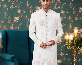 Könnte beinhalten: Eine weiße, langärmelige Kurta mit perlmuttartigen Verzierungen und Blumenstickereien. Die Kurta hat einen Mandarinkragen und einen Knopfverschluss. Das Model trägt eine weiße Hose und weiße Schuhe.