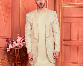 Puede incluir: Un conjunto de kurta y pijama beige con una chaqueta a juego. La chaqueta tiene un cuello chal y está bordada con hilo dorado. La kurta tiene mangas largas y cierre de botón. El pantalón de pijama es liso y tiene una pierna recta. El hombre lleva zapatos blancos.