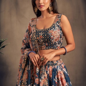 May include: A blue and white floral print lehenga with gold embroidery. The lehenga has a matching blue sleeveless blouse with gold embroidery. The model is wearing gold earrings and a ring.