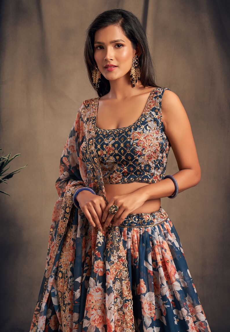 May include: A blue and white floral print lehenga with gold embroidery. The lehenga has a matching blue sleeveless blouse with gold embroidery. The model is wearing gold earrings and a ring.