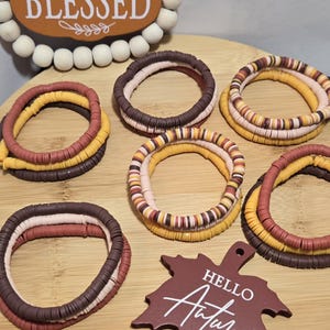 Puede incluir: Varios brazaletes coloridos en tonos marrón, amarillo y rosa están dispuestos sobre una superficie de madera. Un letrero de madera que dice "BLESSED" está en el fondo. También hay un letrero en forma de hoja que dice "HELLO Autumn".