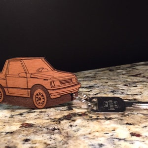 Puede incluir: Un llavero de madera con forma de Suzuki Sidekick marrón. El llavero está unido a una llave plateada.