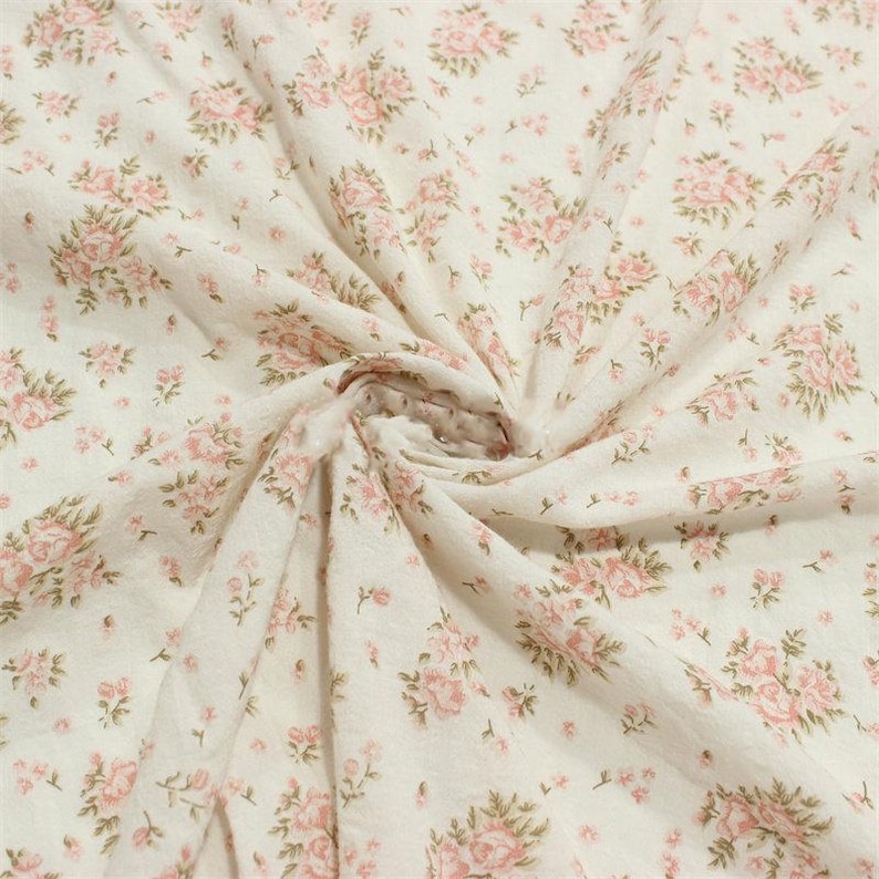 May include: Off-white fabric with a repeating pattern of pink roses and green leaves. The fabric is gathered in the center, creating folds and shadows. The floral design is evenly distributed across the material.