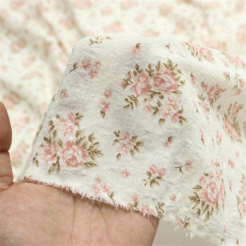 May include: White fabric with a delicate floral pattern featuring pink roses and green leaves. The fabric has a slightly textured appearance and a raw edge, suggesting it may be used for crafting or sewing projects.