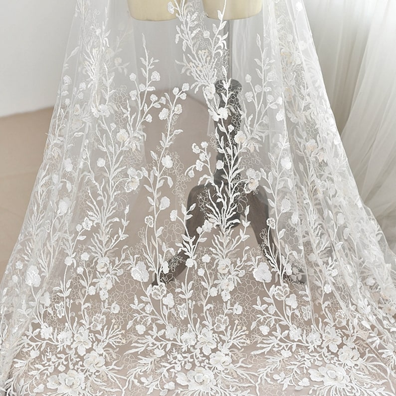 May include: White floral lace fabric with delicate embroidery. The lace features a repeating pattern of small flowers and leaves.
